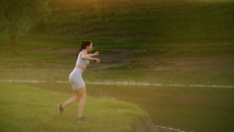 A-woman-trains-on-the-shore-of-a-lake-in-a-park-in-the-sunlight-of-sunset-light.-Cross-lunges-back-with-a-jump.